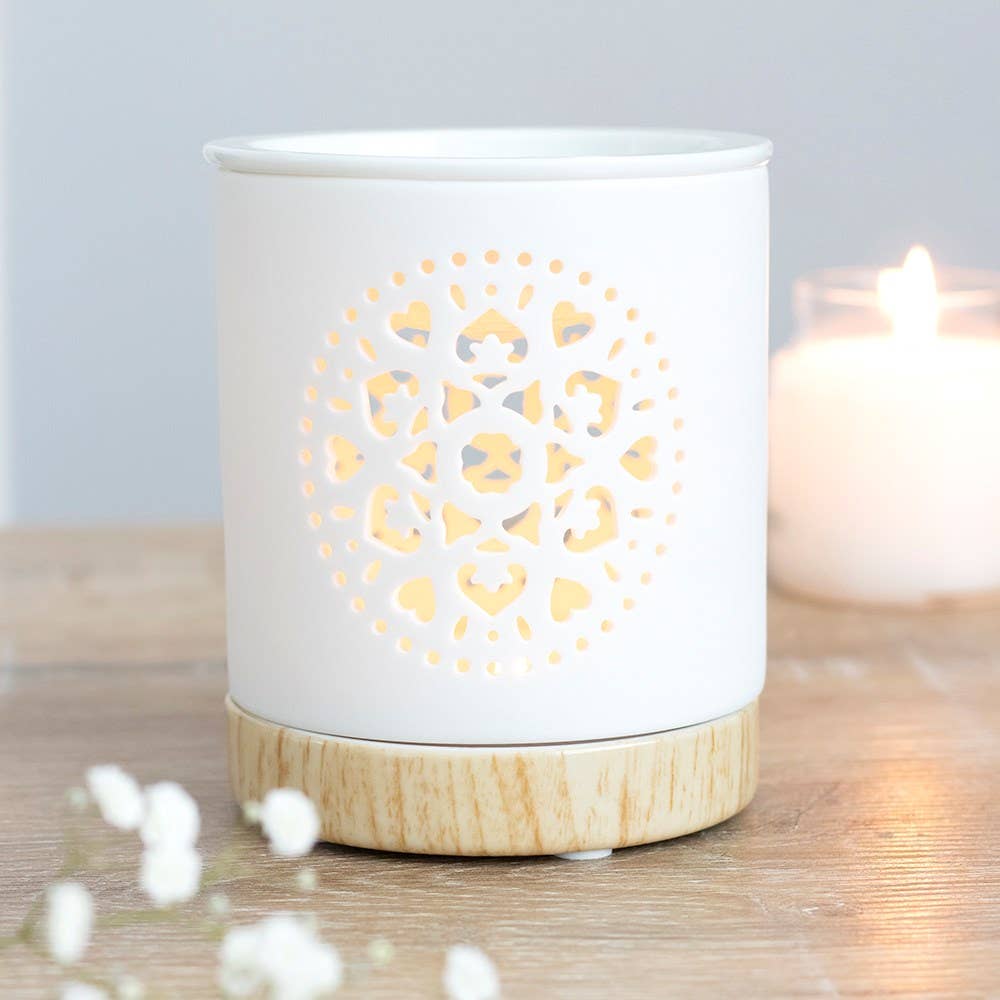 A white ceramic oil burner with a geometric mandala cutout design on the lid, placed on a wooden surface with a candle inside.