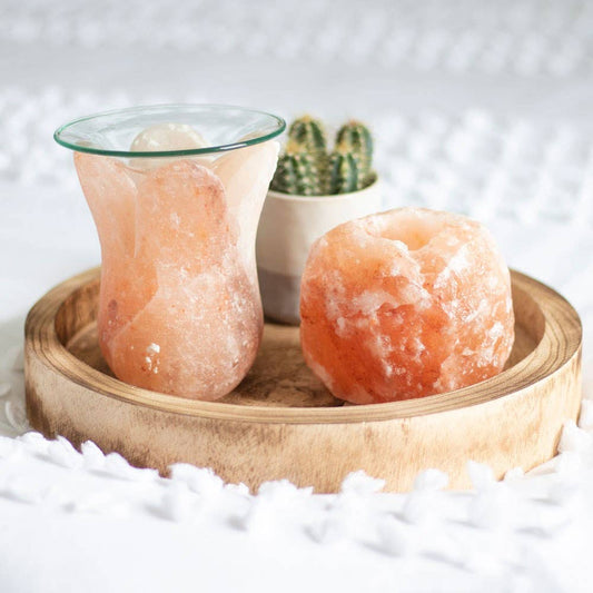 A pink Himalayan salt candle holder in the shape of a rounded bowl with a single tealight candle placed in the center, on a white textured surface with a cactus in the background.