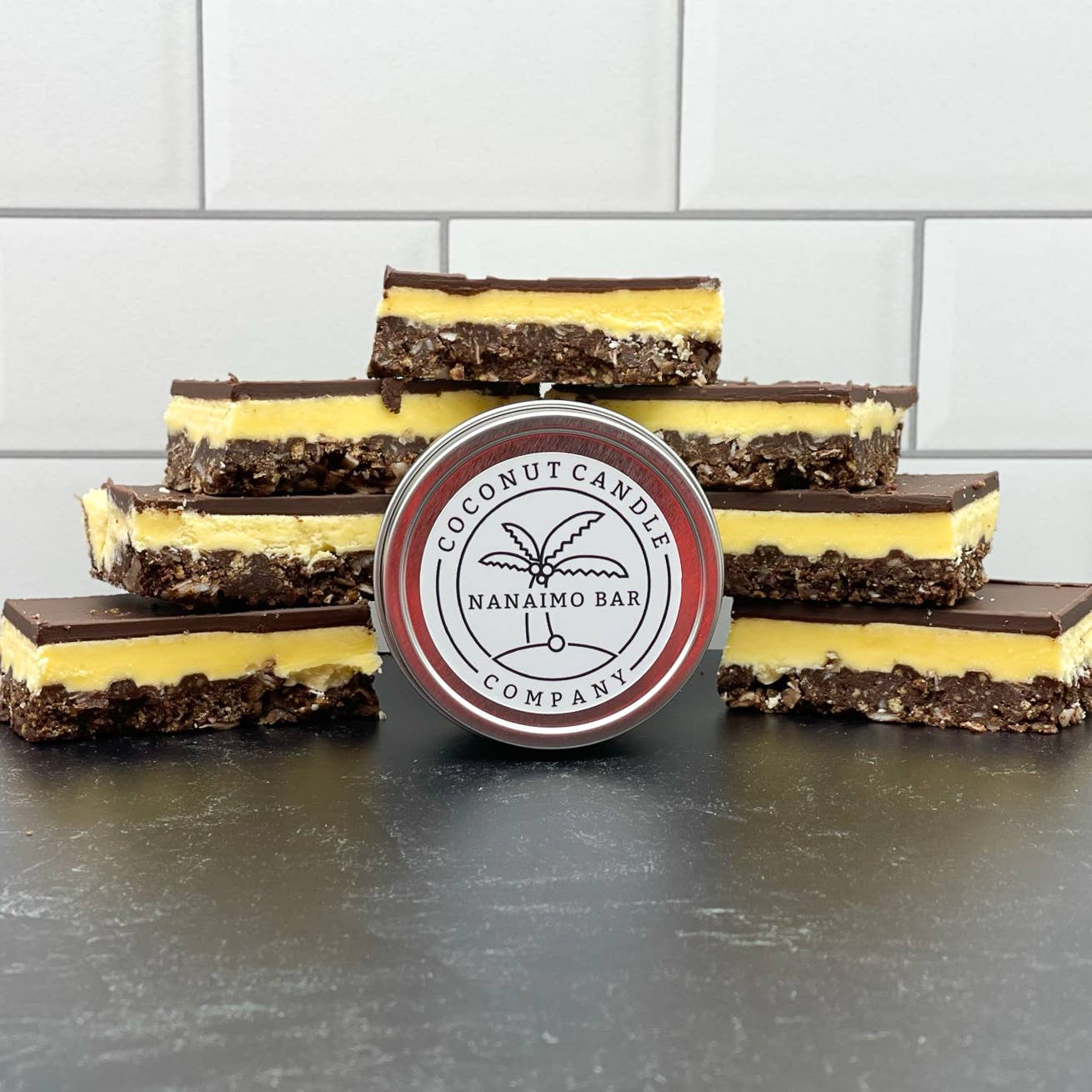A series of Nanaimo bars with a coconut candle company label in the foreground, arranged on a dark surface with a white tiled background.