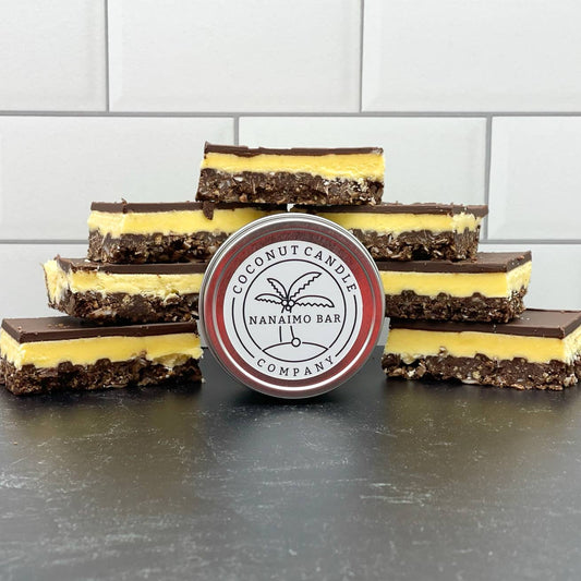A series of Nanaimo bars with a coconut candle company label in the foreground, arranged on a dark surface with a white tiled background.