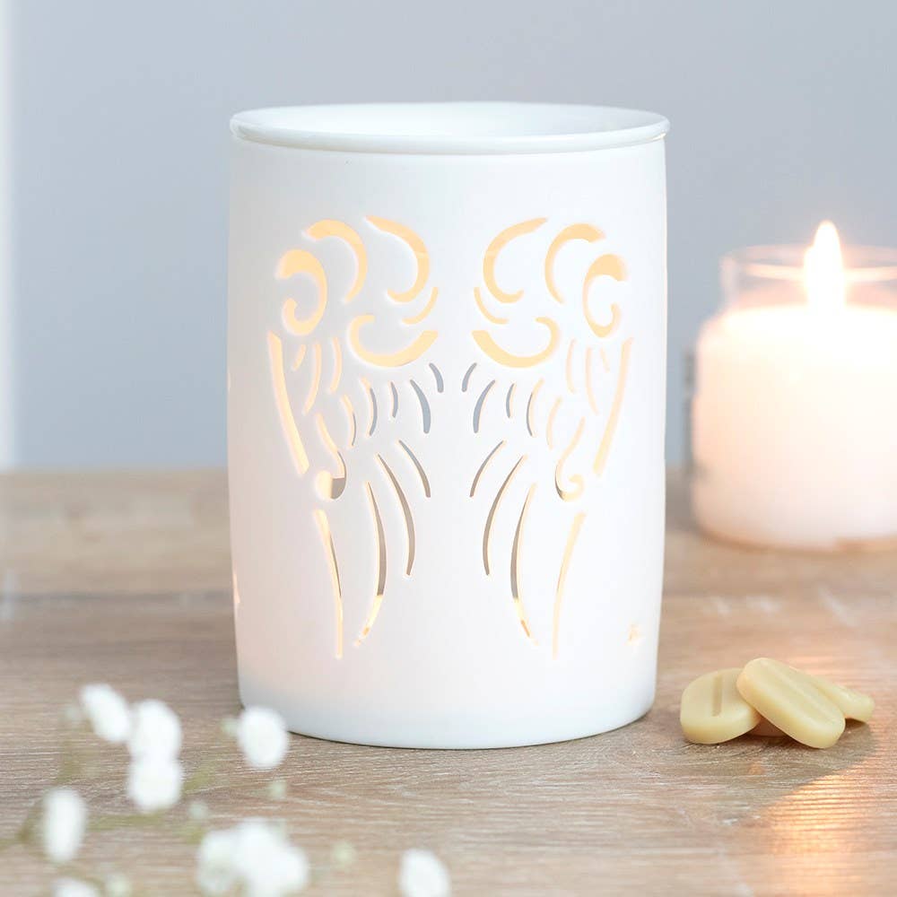 A white ceramic oil burner with cutout angel wings design, accompanied by a lit tealight inside, placed on a wooden surface with some dried flowers nearby.